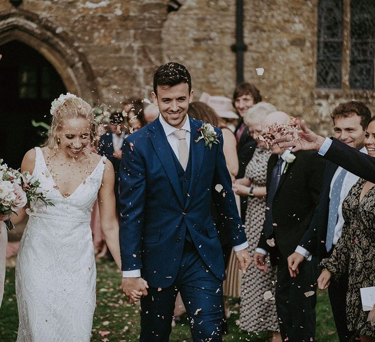 Bride in lace wedding dress and groom in three-piece navy blue suit and beige tie have their confetti exit