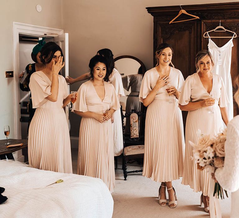 Bridesmaids in pale pink dresses with butterfly sleeves and pleated skirts see the bride in her dress for the first time