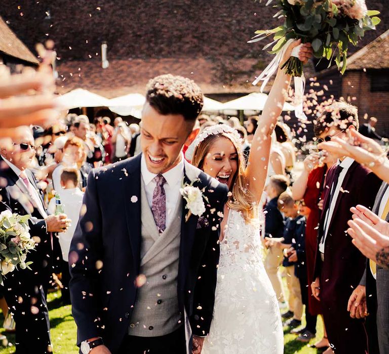 Bride wearing lace wedding dress and white headband walks with groom in three piece suit through confetti 