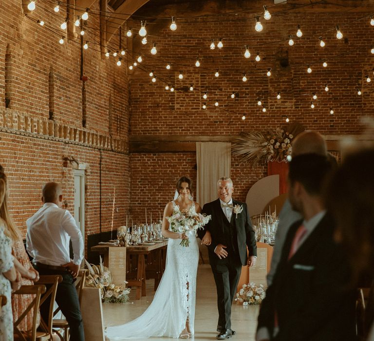 Father of the bride in black tie with gold bow tie walks the bride down the aisle in lace wedding dress for barn wedding with rustic decor 