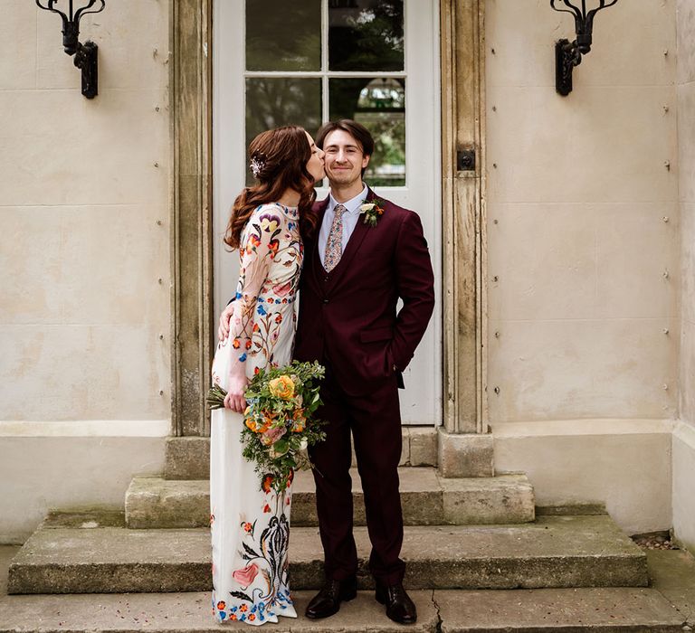 Bride in colourful wedding dress kisses the groom on the cheek wearing burgundy suit 