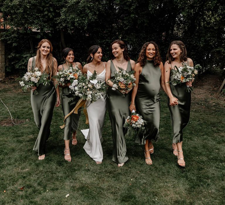 Bridal party in sage green satin bridesmaid dresses holding white and orange flower bouquets with green foliage 