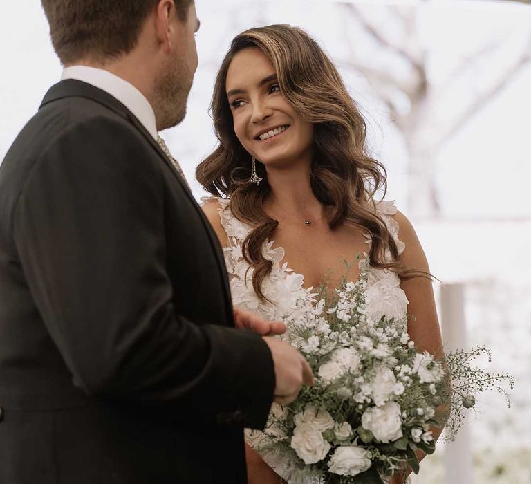 Bride holds white flower wedding bouquet with nude wedding nails and gold bracelet for classic marquee wedding as she stands at the altar with the groom 