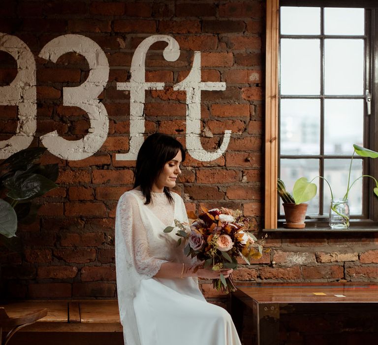 Bride wears wedding cape and sits atop wooden table whilst looking down toward floral bouquet filled with Autumnal coloured blooms