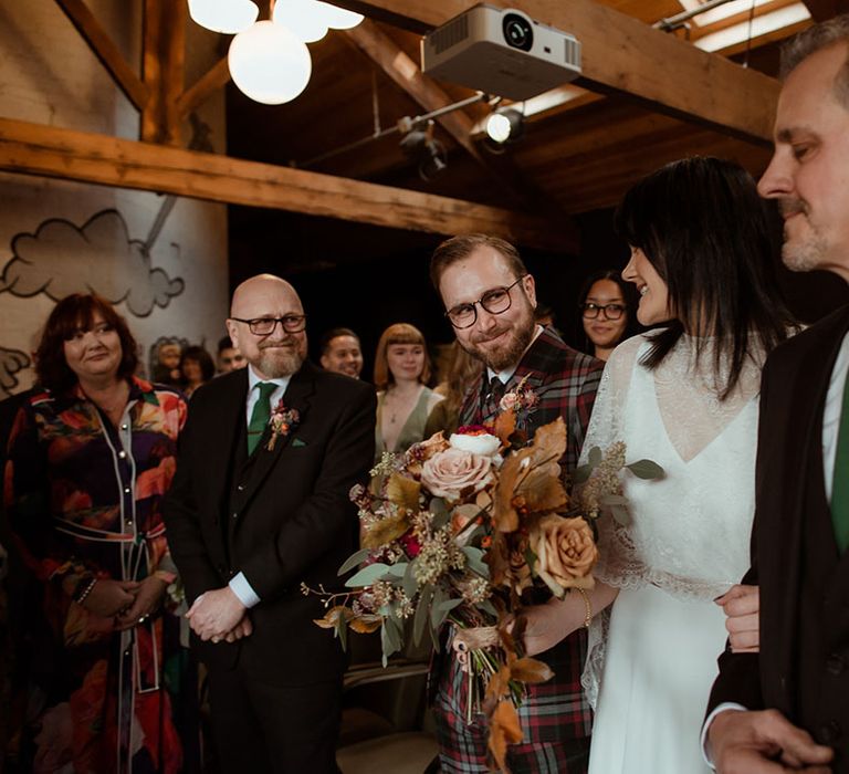 Wedding guests smile at one another at The Chimney House as bride holds Autumnal floral bouquet 