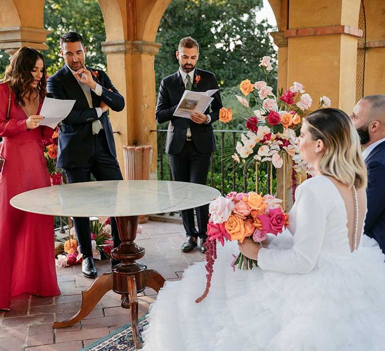 Bride & groom kneel outdoors during wedding ceremony in Florence in front of bright floral bouquets 