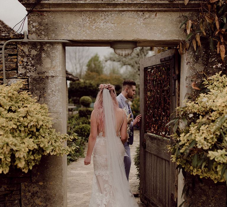 Bride wearing lace wedding dress from Jane Hill walks through rustic door outdoors to intimate garden for first dance with her groom