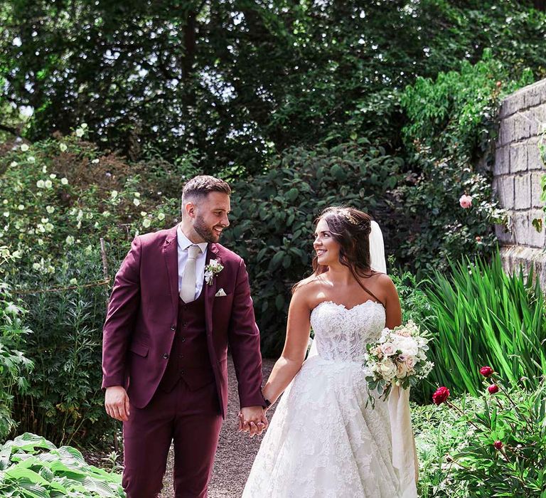 Bride in a strapless lace wedding dress holding hands with there groom in a burgundy coloured wedding suit in the gardens at East Riddlesden Hall