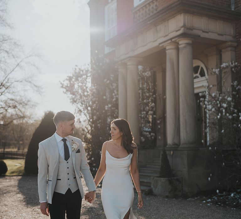 Bride in cowl neck wedding drew with front slit walking holding hands with groom in a grey suit jacket and blue tie 