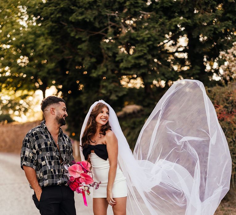 Bride wears white mini dress with black bow and throws out her veil beside her as her groom watches on 