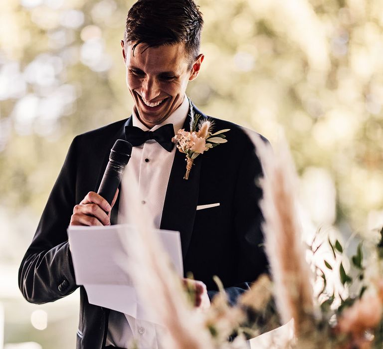 Groom wearing black tie and pampas grass buttonhole tied with brown string gives speech during reception