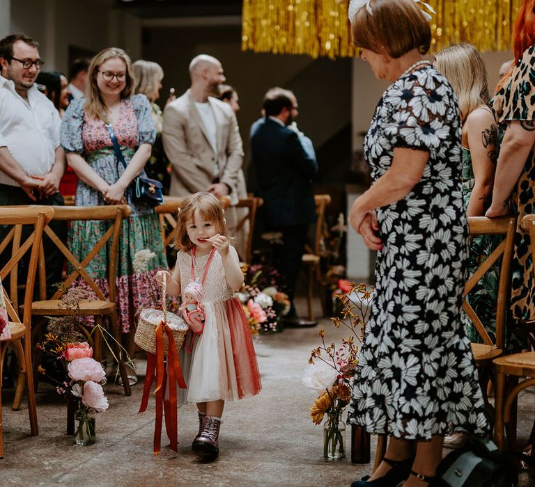 Flower girl wears pink dress and holds wicker basket tied with orange ribbon as she walks down the aisle