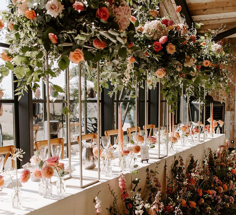 Brightly coloured floral installations line banquet tables with white tablecloth for barn wedding reception 