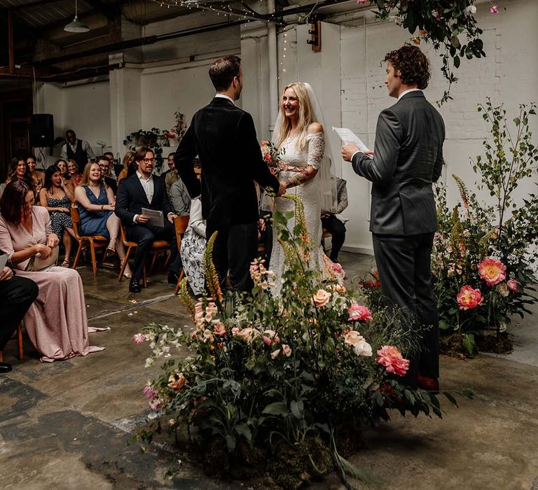 Bride and groom smiling at each other at the alter with large foliage arch decorations with colourful floral details and fairy lights