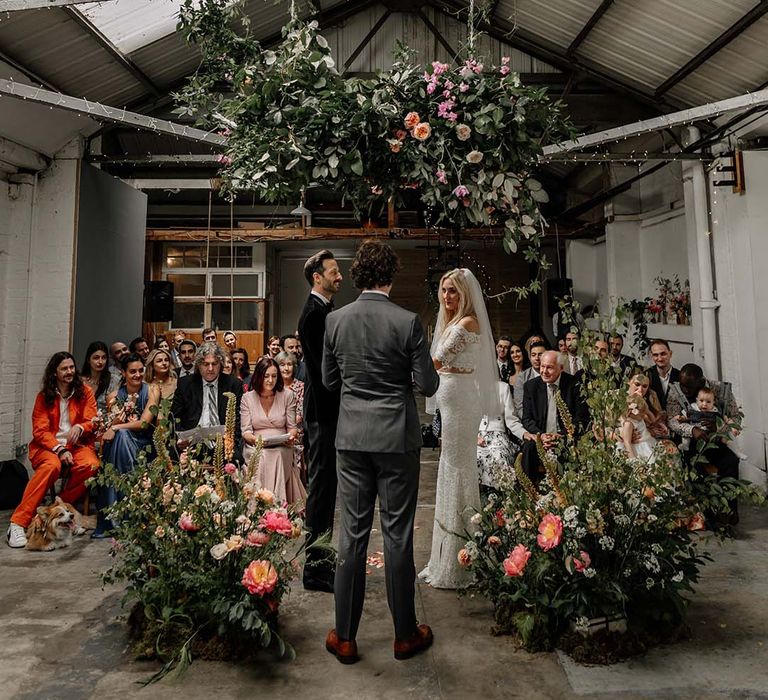 Bride and groom embracing at alter with large foliage arch decorations with colourful floral details and fairy lights