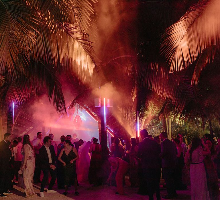 Wedding guests party outdoors in the jungle surrounded by pink lighting and smoke