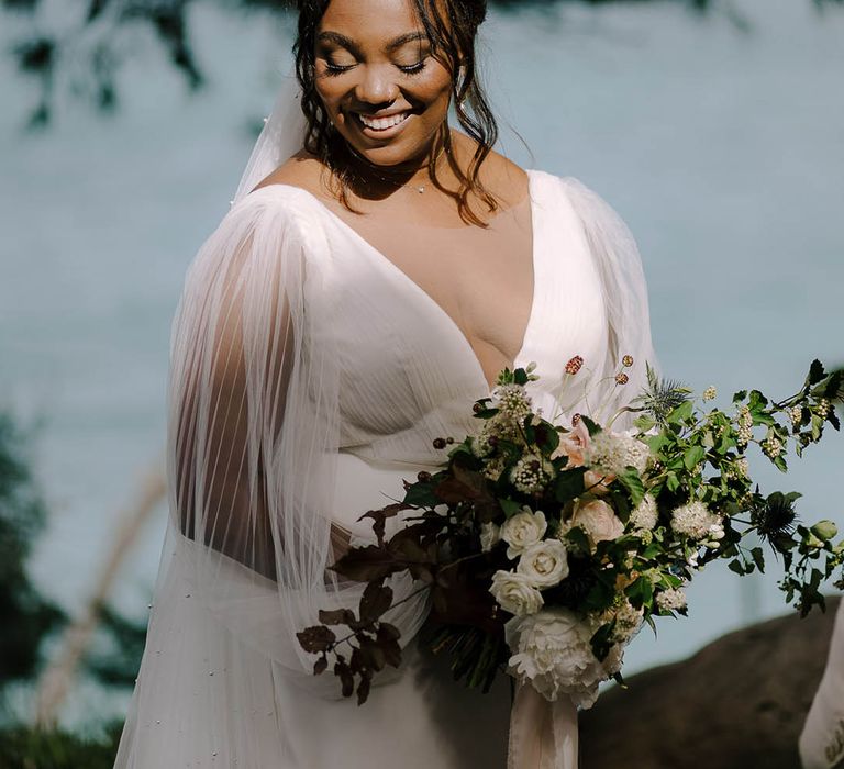 Bride shows off sheer-sleeved wedding dress in the sunshine with rose and wildflower bouquet