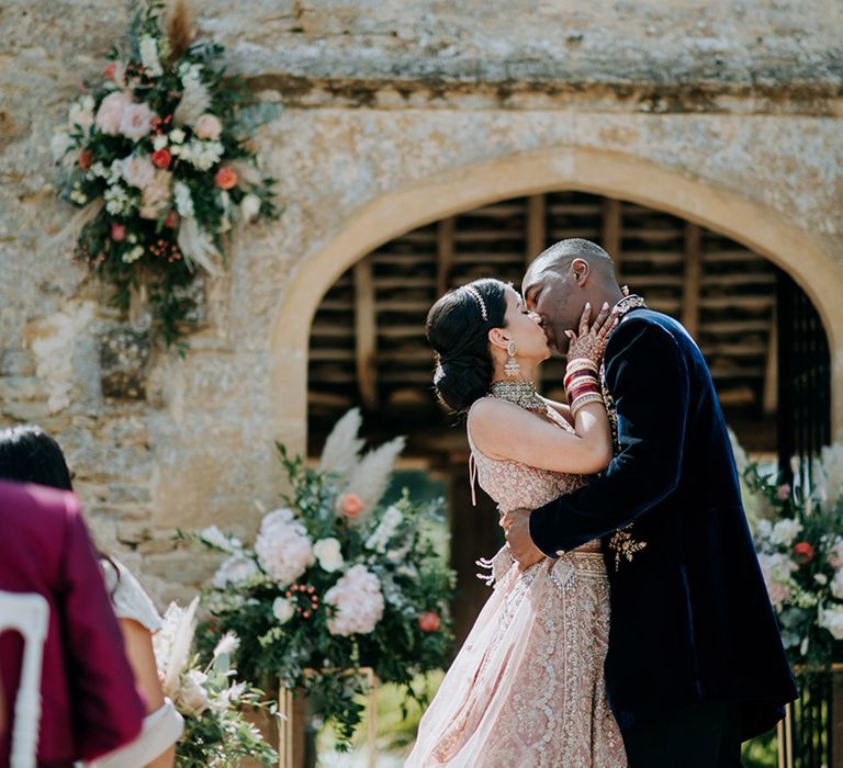 Multicultural Bride and Groom kiss at floral decorated alter at Cotswolds venue Caswell House