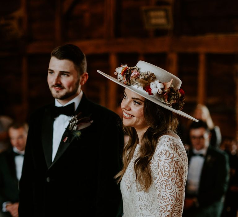 Bride wearing a laser cut lace wedding dress with bell sleeves and wedding hat standing with the groom at the altar 