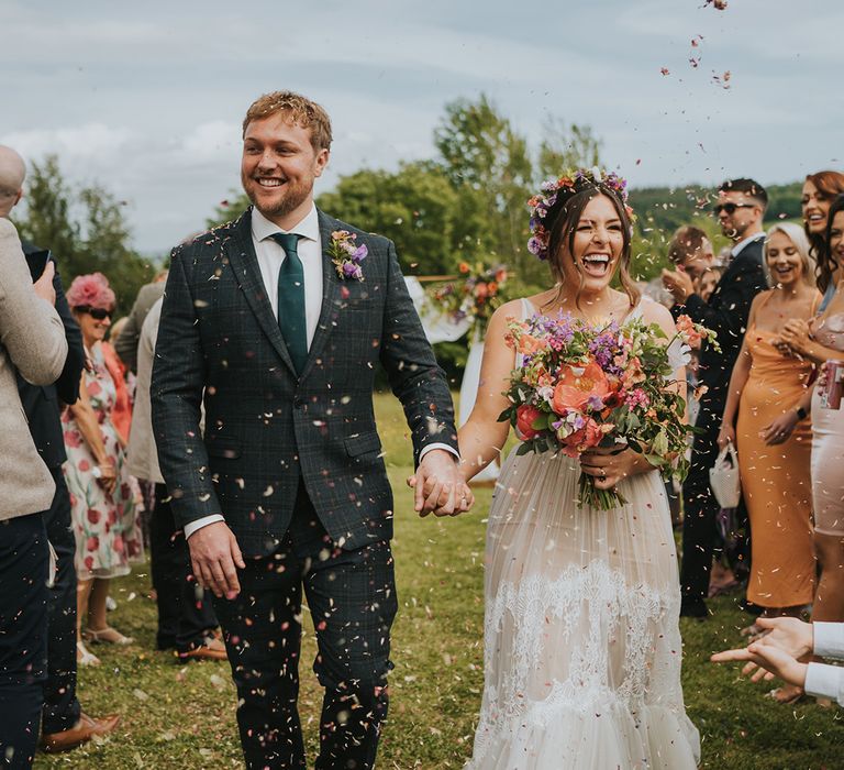 Bride carrying colourful bouquet walks through colourful confetti exit with her groom