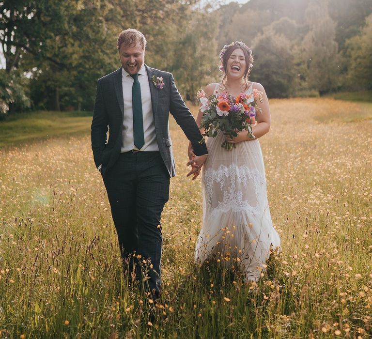 Bride holding colourful bouquet walks through fields with her groom during golden hour couples portraits 