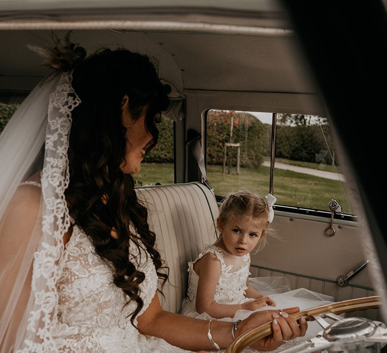 Bride in a tulle and lace wedding dress wearing a lace edge veil sitting in the wedding car with the flower girl in white dress and white hair bow accessory