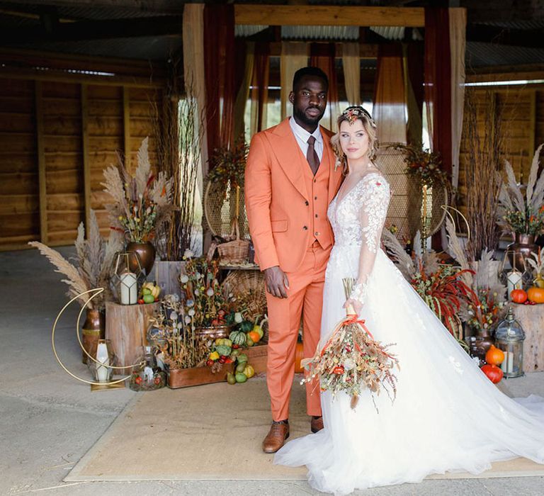 Groom in burnt orange rust coloured suit with brown tie, pocket square, brown accessories and dried flower, rose and pampas grass boutonniere and bride in long sleeved v neck soft ivory dress with lace floral detailing on the bodice and sheer sleeves 