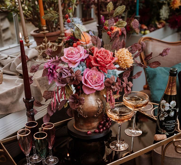 Black glass and gold drinks cart with champagne glasses and cocktail glasses, a bottle of champagne, and a single tier iced wedding cake in the style of an aged golden vase illed with handmade wafer paper florals