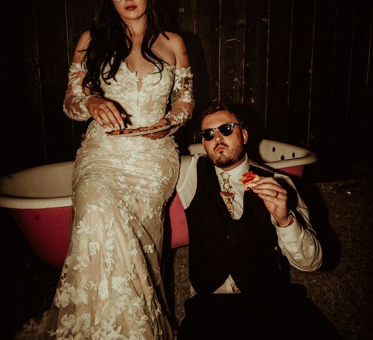 Bride in off the shoulder Abigail Westrup bridal gown with long, sheer sleeve and lace detailing wearing cat eye bejewelled sunglasses sitting on the edge of a bathtub with groom in black waistcoat and flower patterned tie wearing Raybans sunglasses both eating pizza 