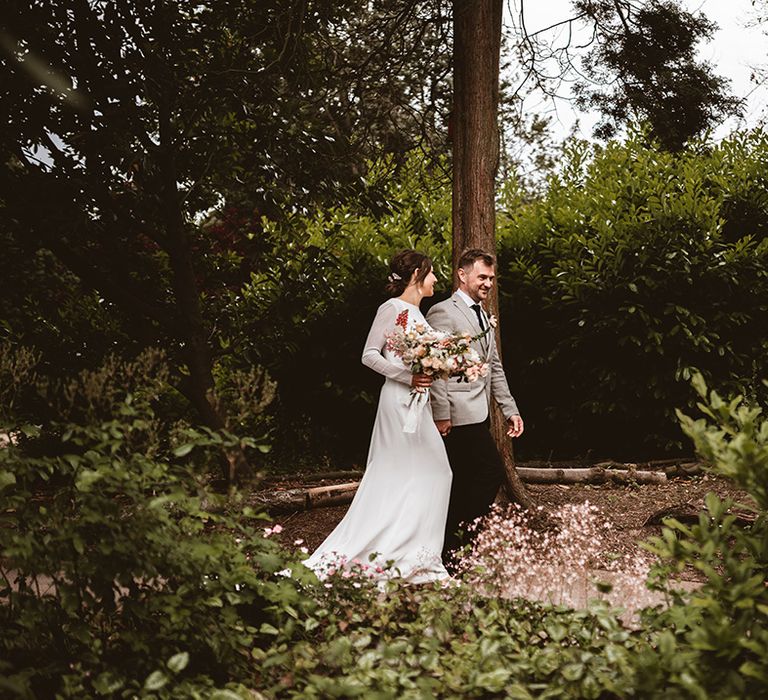 Groom in black suit trousers, grey blazer and light pink rose boutonniere and bride in long satin wedding dress with bridal cover up semi sheer crop top walking in the wedding venue gardens