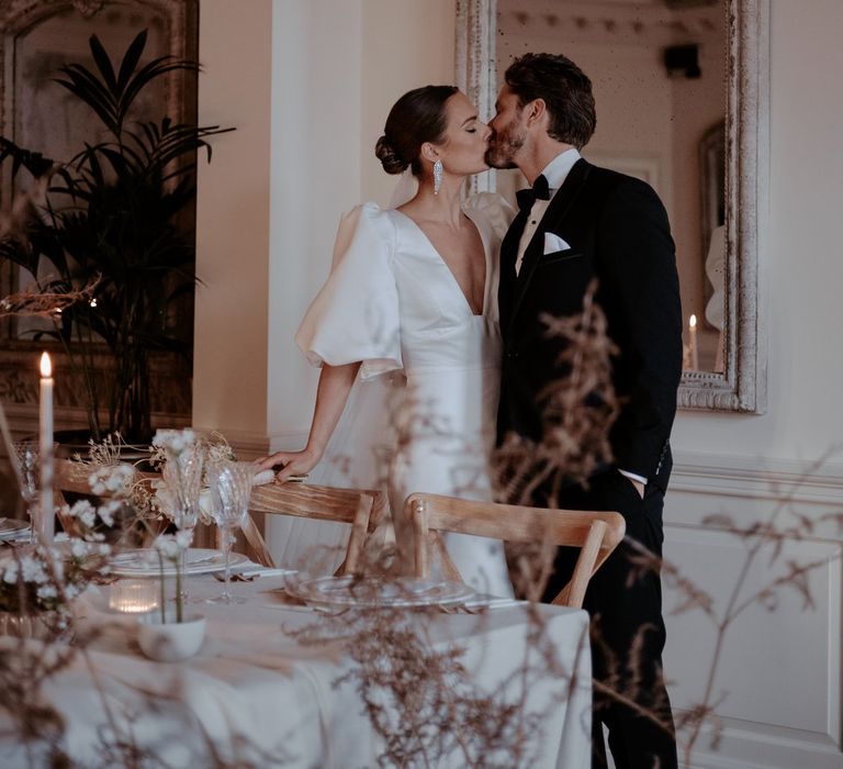 Bride in v neck puff sleeve wedding dress with front slit and church length veil and groom in classic black tux kissing in reception room of Eaves Hall with large dried flower arrangements and tapered candles 