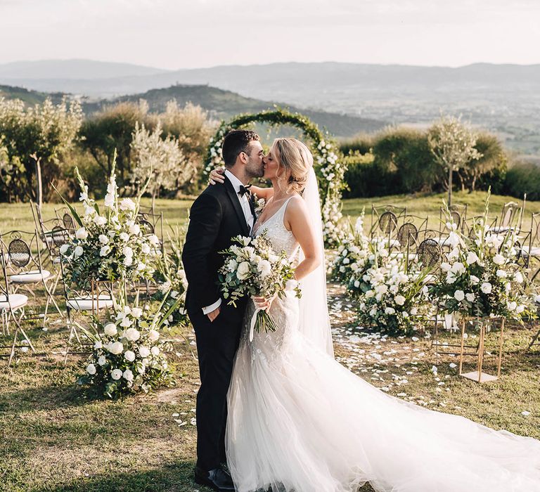 Bride in fitted wedding dress and groom in black-tie kiss outdoors during Italian reception 