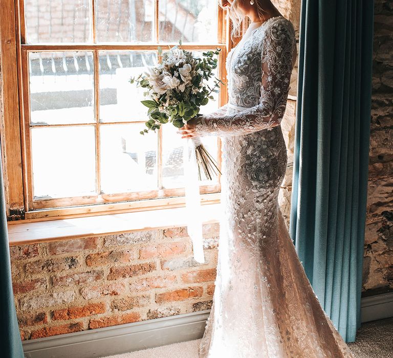 Bride in 3D lace wedding dress with long sleeve and train with pearl earrings holding white flower bouquet tied with white ribbon 