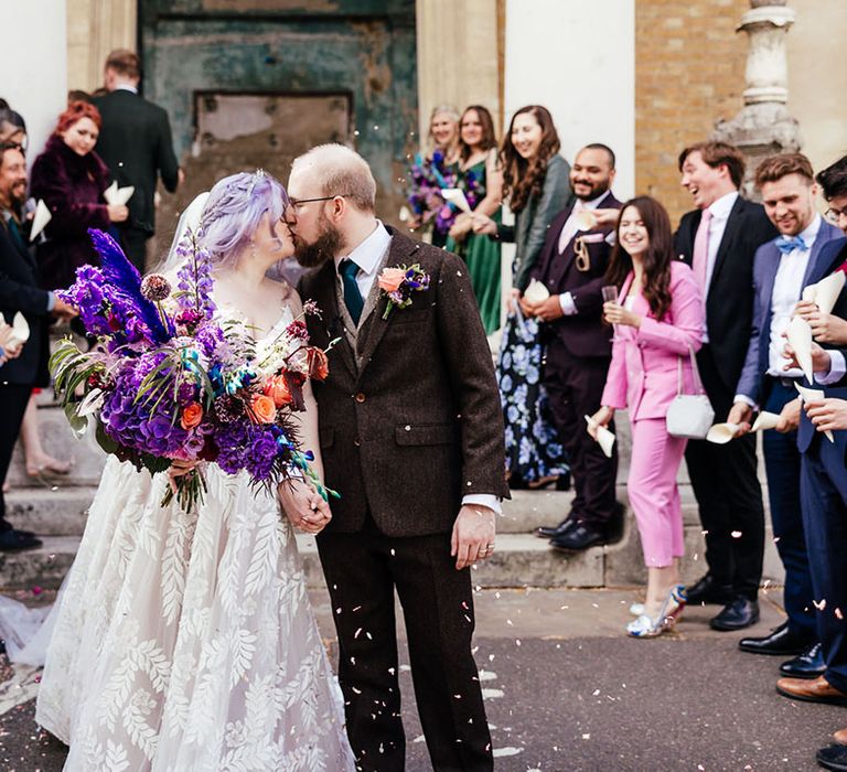 Bride wearing leaf appliqué sheer Evie Young bridal gown and cathedral length veil holding bright purple bridal bouquet with purple hydrangeas, pink garden roses, purple orchids, lilac carnations, dried flowers and peacock blue feathers kissing groom in mocha brown textured suit with deep green tie and pink and purple boutonniere at Asylum Chapel London wedding venue   