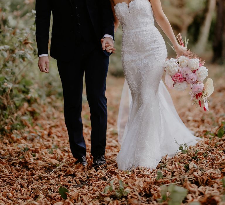 Bride in strapless mermaid wedding dress holding an oversized pink and white bouquet walking with the groom in a blue suit 