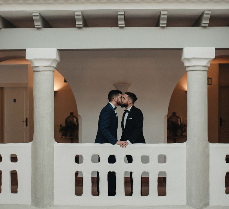 Groom in tuxedos and bow ties kissing on a balcony at their destination wedding 