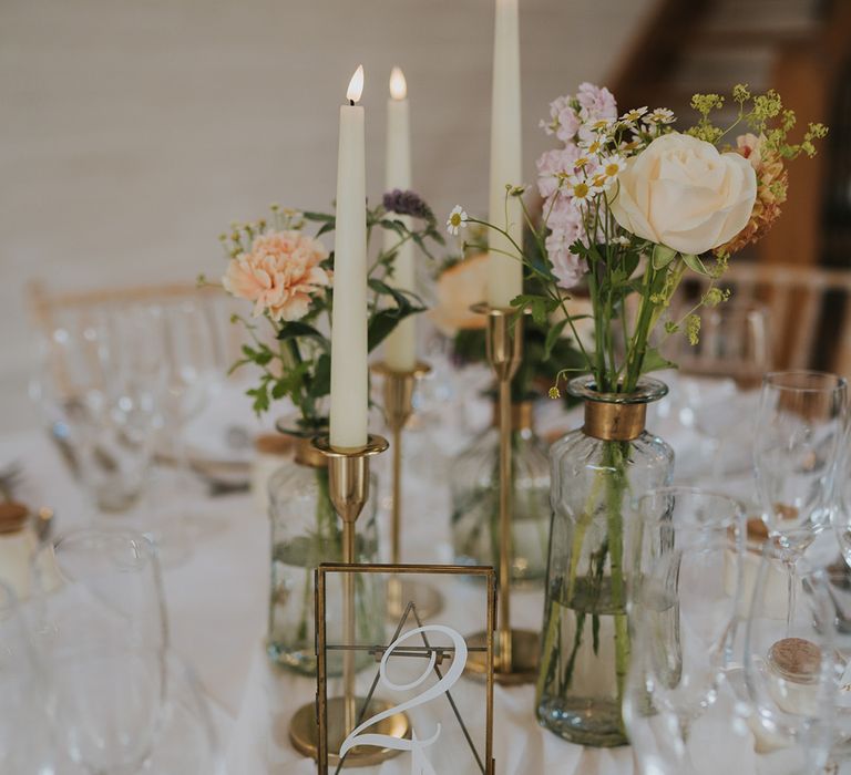 Clear glass table number sign on gold photo frame with white taper candles in gold candle holders and pastel wedding flowers in bud vases 