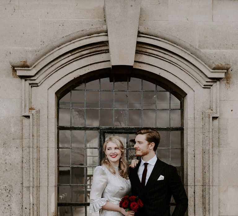 The bride wearing red lipstick, pearl earrings and silver wedding dress holding red rose bouquet with the groom in a pinstriped suit 