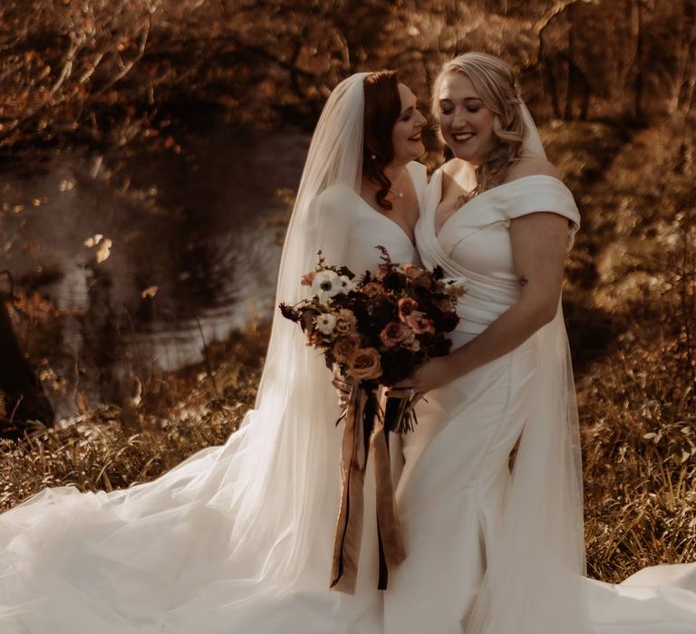 Bride in long sleeve deep v wedding dress and church length veil and bride in satin short sleeve deep v wedding dress and church length veil standing by the river at Hidden River Cabins