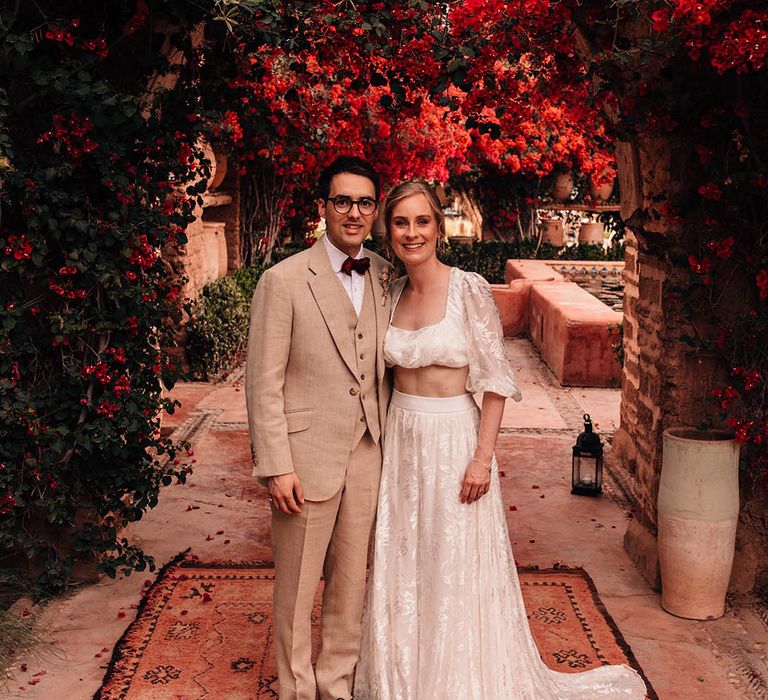 Groom in cream three piece suit with red bow tie and dried flower buttonhole with the bride in modern two piece set 