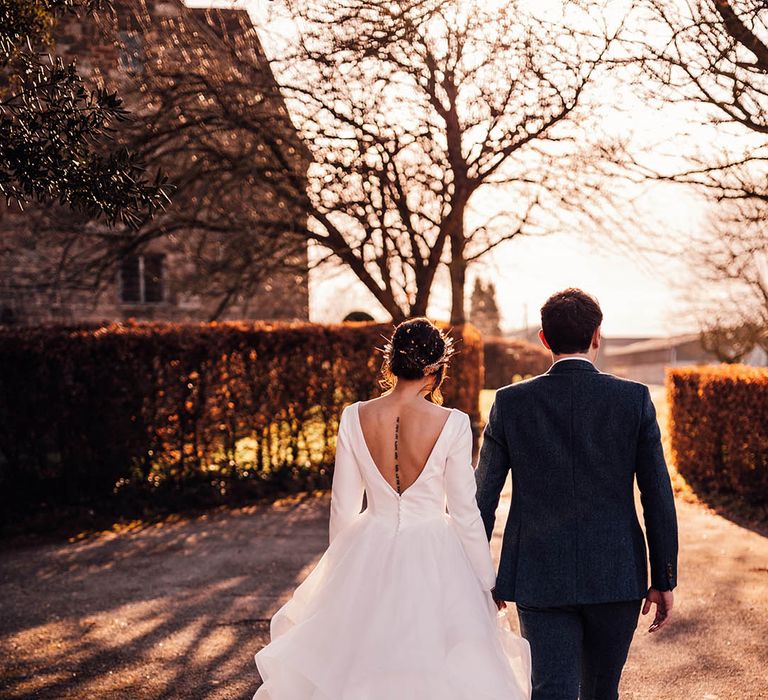 Bride in low back dress with full princess skirt walking around their wedding venue with groom in navy suit 