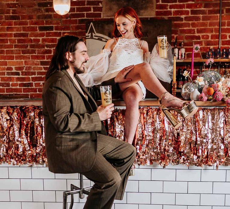 Groom in deep brown suit with off-white t-shirt and vans sitting on bar stool next to wedding bar with bride in 70s-inspired boatneck short lace wedding dress with detachable tulle puff sleeves and platform gold wedding heels sat on top of the bar with rose gold and orange metallic streamers 