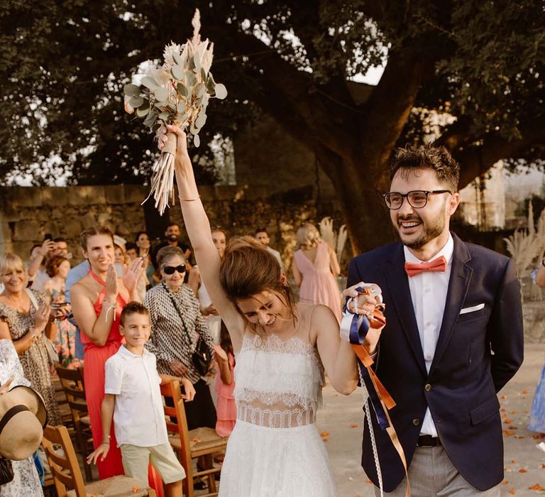 confetti exit at greece destination wedding with boho bride in a lace wedding dress waving her pampas grass bouquet in the air holding hands with her groom in a navy blazer and orange bow tie