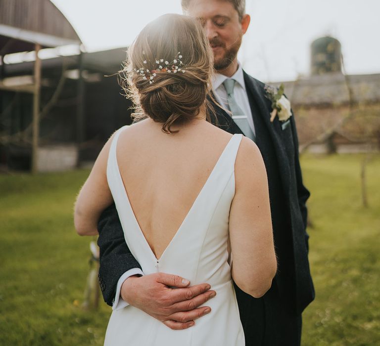 Bride wearing a classic low back wedding dress with her hair in an updo styled with gypsophila being embraced by the groom 