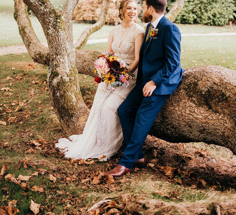 Groom in blue suit with bride sitting on log at rural rustic grounds of country house wedding venue 