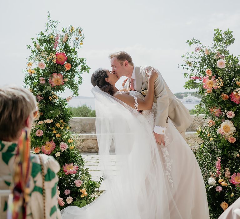 Groom in linen suit dips bride for first kiss at altar decorated with flower columns at destination wedding in Hvar Croatia 