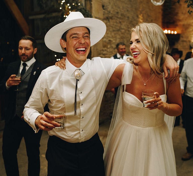 Groom in white shirt and white cowboy hat with traditional bolo tie laughing with the bride in sparkly wedding dress 