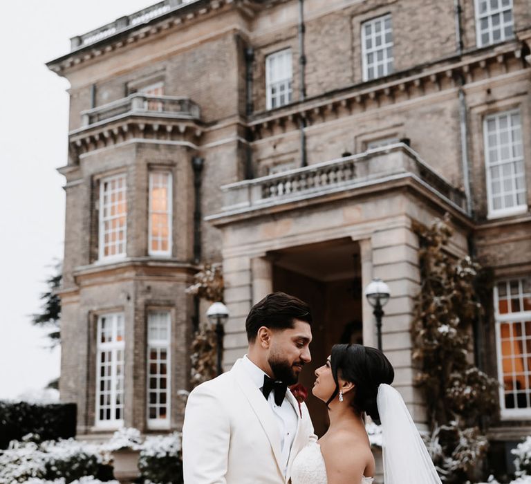 Groom in white tuxedo with bride in fitted lace mermaid wedding dress in front of Hedsor House 