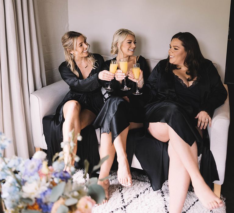 Bridesmaids in black satin robes sit together as they get ready together drinking mimosas 