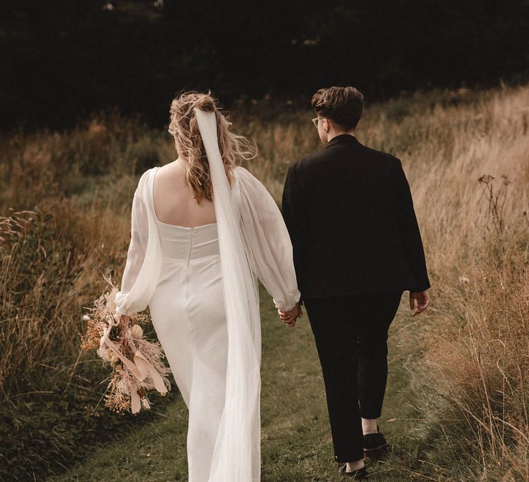 Bride wearing wedding veil and gown walking along with bride in brown suit around the wedding venue in Wales 